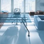 Nowoczesne ergonomiczne biuro ze szklanym biurkiem, niebieską sofą i minimalistycznymi meblami w odcieniach brązu.