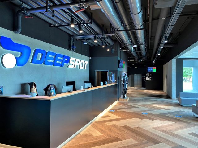Projekt nowoczesnej recepcji Deepspot z industrialnym sufitem, drewnianą podłogą i ergonomicznym stanowiskiem.