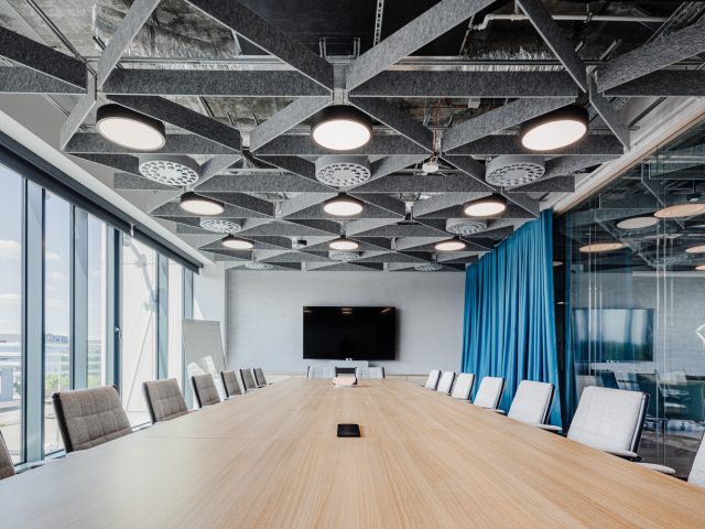 Nowoczesna sala konferencyjna z drewnianym stołem, ergonomicznymi krzesłami, akustycznym sufitem i dużymi oknami.