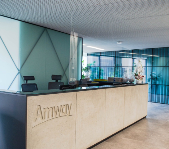 Elegancka recepcja biura Amway z beżowym blatem, minimalistycznymi szklanymi ściankami i szarym sufitem.
