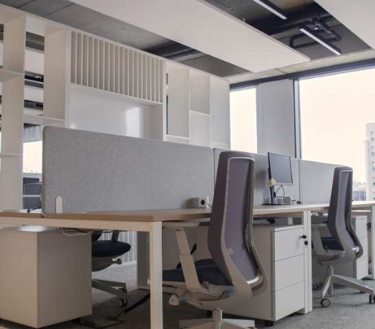 Ergonomiczne stanowiska pracy w nowoczesnym biurze open space, jasne meble i szare panele akustyczne.