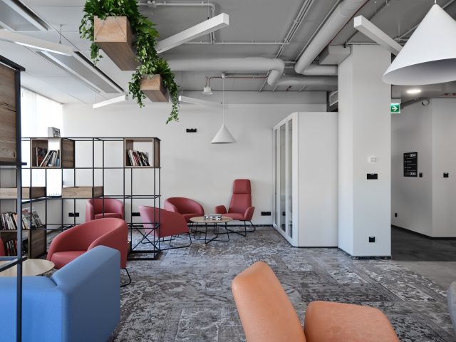 Ergonomiczna strefa współpracy w biurze z czerwonymi fotelami, regałem, zielonymi roślinami i akustyczną budką.