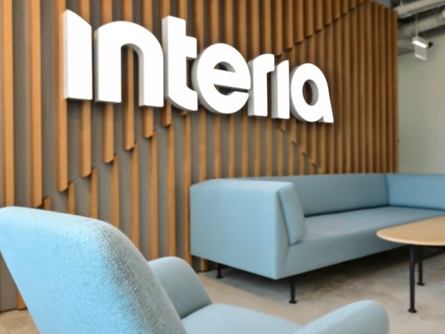 Nowoczesna aranżacja recepcji biurowej z logo Interia, jasnoniebieskimi meblami wypoczynkowymi i drewnianymi panelami.